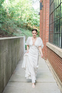 A person in a wedding dress walking down a sidewalk and holding the bottom of their dress with one hand