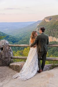A bride and groom stand at an overlook looking out enjoying the views. Their backs are facing the camera with the grooms arm wrapped around the bride. The bride is wearing a lace white wedding gown with a train and the groom is in an olive green suit. There are rock and tree covered mountains in the distance with the sky turning pink from the sunset.