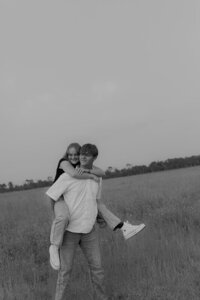 Black and white photo of a man is giving his girlfriend a piggy back ride through a grass field during a couples shoot in Rexburg, Idaho.
