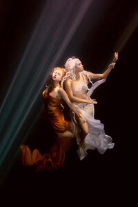 Two women dressed in flowing golden and white gowns gracefully floating underwater, illuminated by beams of ethereal light. One woman, with red hair and golden attire, closes her eyes in serene elegance, while the other, with platinum hair and white attire, reaches upward with poised grace. Their movements convey a dreamlike beauty in this captivating underwater scene.