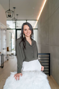 Brand Headshot of Nutritionist wearing olive top posing in bright modern home leaning on furry chair 