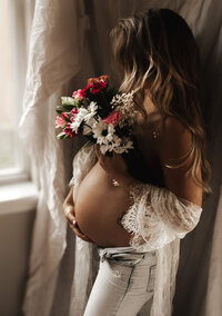 Pregnant woman holding a colorful bouquet near a window in soft lace, captured during a cozy indoor maternity session in the Pacific Northwest.