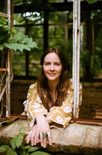 A woman with long brown hair and a yellow patterned top leans on a brick windowsill, smiling, with greenery and rustic window frames in the background—a perfect scene for a Showit designer or brand and web design inspiration.