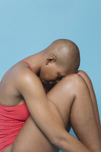 A black woman with a shaved head sits on the ground, head against her knees. Her eyes are closed, with a solemn expression.
