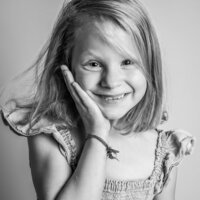 A young girl with blonde hair smiles sweetly at the camera, resting her face in one hand. Her hair is slightly tousled, and she wears a ruffled dress.