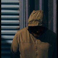 Figure in a brown hooded jacket standing against a blue corrugated metal wall, with their face mostly obscured by shadows.