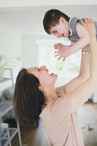 Kaya lifting her laughing child Luca into the air while smiling in a bright room.