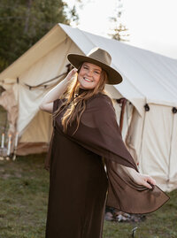 Sydney Breann, a Montana-based elopement photographer, smiles while posing in front of a canvas tent during golden hour at a lakeside campsite.