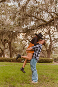 A playful couples session in Savannah, Georgia, featuring a man lifting his partner into the air as they smile and kiss under moss-draped oak trees in a historic southern park setting.