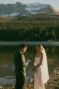 groom reading from paper to bride in front of lake and mountains