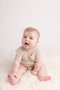 6 Month old baby sitting by herself in a cute beige knit romper, sitting on a beige carpet. she is smiling towards the camera and leaning over to touch one of her ankles. She has the sweetest baby rolls.