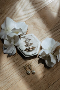 A brides engagement and wedding rings along with a groom's ring in a hexagon ring box surrounded by florals and the bride's earrings.