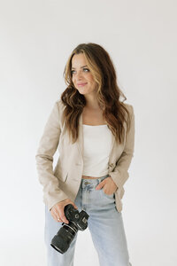 Branding photo for photographer, a blonde woman wearing jeans, white top, and olive green blazer while holding her phone and smiling at it