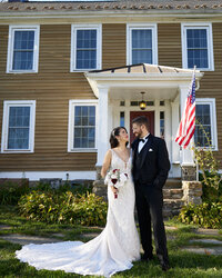 A bride and groom stand smiling at each other in front of a tan house with white trim and an American flag. Captured by a talented NJ wedding photographer, the bride wears a white lace gown with a bouquet; the groom wears a black tuxedo.