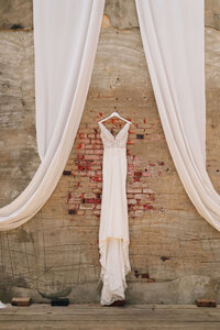 A white wedding dress hangs on a wooden hanger against a brick and concrete wall, framed by flowing white drapes on either side.