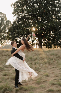A groom spins his bride while holding her up, showing beautiful motion in her dress and hair. Behind them are tall trees with the sun peeking through.