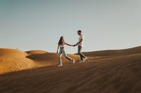 A couple walks hand in hand across sand dunes during their engagement session in Yuma, Arizona. Both are dressed casually in jeans and t-shirts, enjoying the expansive desert landscape around them.