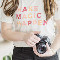 woman holding a camera and wearing a t-shirt that says "make magic happen"