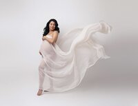 Pregnant woman wearing a white silk fabric and a fan blowing the fabric with her posed.