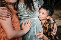 close up of dark haired dark eyed boy's stare as he holds his mothers side.  She is wearing a silvery blue dress and has her daugher in an orange lace dress holding her other side.  The only visible face is the boy. He is wearing yellow and black plaid.
