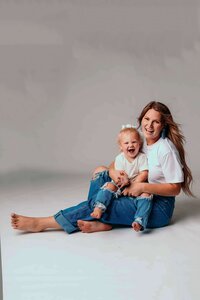 A young mom with long blond hair sits in the studio floor of Mobile AL Family Photographer with her toddler daughter in her lap. They are wearing matching jeans and shirts, and are smiling at the camera.