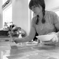 Artist, Meg Pohlod, applying adhesive to a surface with a wide brush while holding a bowl, preparing materials for a collagraph plate in a studio setting.
