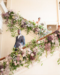 A groom waits on an elegant staircase decorated with elaborate florals for a first look with his bride