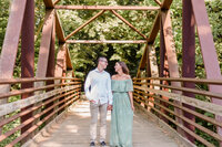 A couple poses for the camera looking at one another while standing in the middle of an iron bridge.