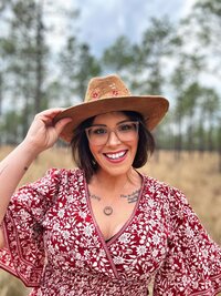 Portrait of Nikki Rinc, founder of Salt + Heart Photography, smiling in a floral dress and hat during a branding session in a natural Florida field
