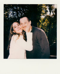 Polaroid photo of an engaged couple sharing pizza on the floor of a styled indoor set with tropical leaves and disco balls — captured by a film wedding photographer during an engagement session.