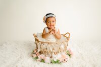one year old little girl from charlotte, NC dressed in cream on a cream background smiles with her hands by her face while sitting in a boho style basket with flowers 