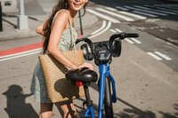 A joyful woman is captured in a sunlit urban setting, leaning on a blue city bike.
