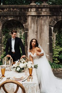 Bride leaning on a table with elegant vintage place settings and groom leaning behind her gazing.