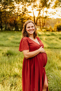 glowing mom to be in a red dress smiling while holding her bump during maternity photo session