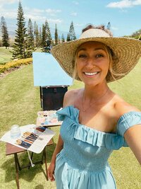 an artist takes a selfie in front of her live wedding painting set up of easel, canvas, paints, and brushes  at the grassy ceremony site