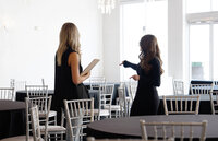 Two women discuss event setup inside SoHo63, a Chandler wedding and event venue. Surrounded by silver chiavari chairs and black table linens, one holds a clipboard while the other gestures toward the bright, modern space during a planning walkthrough.