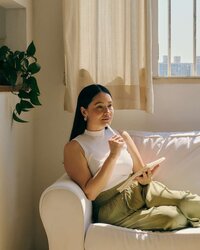 Woman with long hair sitting on a white sofa next to a bright window, holding a tablet and pen. She appears to be thinking or pondering, possibly about custom Showit website design, with sunlight illuminating the room. A plant is visible on a shelf to the left.