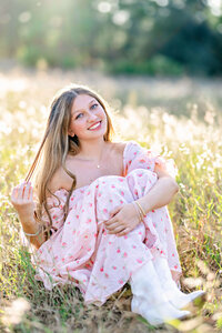 Senior portraits of gorgeous senior girl in Winter Garden, FL. She is modeling a beautiful floral print dress with her long blonde hair while twirling a little piece in one hand situated in a seating position on the ground at Lake Louisa State Park.