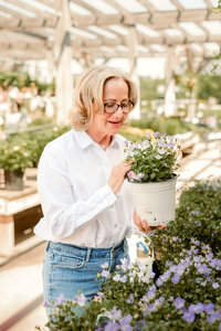 Blonde woman in a white blouse and jeans smiling while holding a lush green potted plant, perfect for an approachable and down-to-earth online dating profile.
