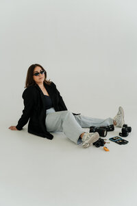 Photographer sitting on the floor in sunglasses and blazer, surrounded by camera gear in studio setting.