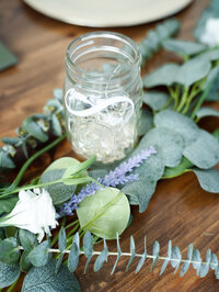 A mason jar filled with twinkle lights is positioned amidst fresh eucalyptus branches along with white and purple flowers on a rich, wooden table. Photo by SAVI Photography - San Diego California Photographer