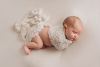 Newborn baby girl laying on side, asleep, with white lace fabric wrapped around her arms and bottom on white fabric backdrop