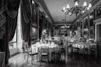 Black and white wedding photo of the grand ballroom at the Goodwood House wedding venue with draped curtains. chandelier and tall floral displays on round tables.
