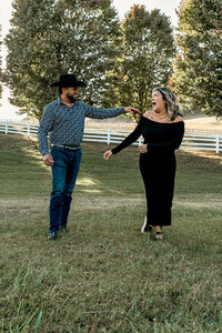 An engaged couple walks outdoors, smiling at each other as they reach out to hold hands. The woman wears a simple black dress, while the man wears a button-up shirt, jeans, and a cowboy hat.