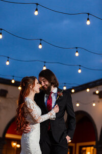 red haired bride in wedding dress nuzzling groom with beard in suit under twinkly lights at dusk at stoney ridge villa wedding venue in azle texas
