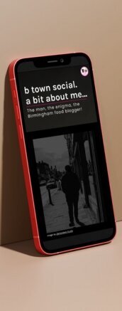 A red smartphone displaying the "About Me" page of the B Town Social website, leaning against a beige surface. The screen shows a black and white image of a person walking down a street with the text, "b town social. a bit about me... The man, the enigma, the Birmingham food blogger!" The overall design is clean and minimalist, focusing on the personal narrative and urban exploration of the blogger.