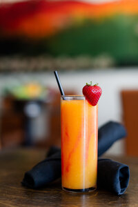 Food photography of an orange and red juice in a tall glass with strawberry garnish and black straw on a wood table with black napkins.