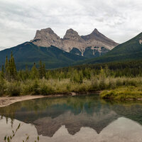 The Three Sisters mountain range in Canmore, Alberta, with lush green forests in the foreground and a reflection of the mountains in nearby water.