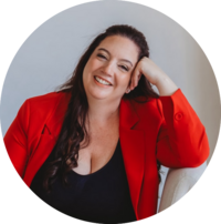 Smiling woman sitting on a cream-colored chair wearing a red blazer and dark jeans, leaning casually with a confident pose.