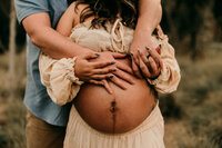 Close-up of a pregnant belly with both parents’ hands gently placed on it during a golden-hour maternity session, featuring a flowing boho dress